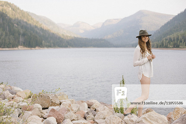 A woman in a hat and white shirt with a basket  by the shore of a mountain lake.