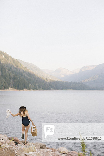 Eine Frau im Badeanzug  die einen Korb am Ufer eines Bergsees entlang trägt.