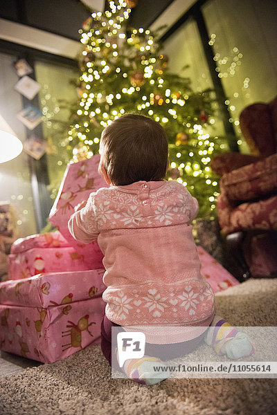 Ein Kleinkind neben einem Haufen Geschenke unter einem Weihnachtsbaum.