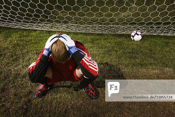 A goal keeper with his head in his hands  and a soccer ball at the back of the goal net.