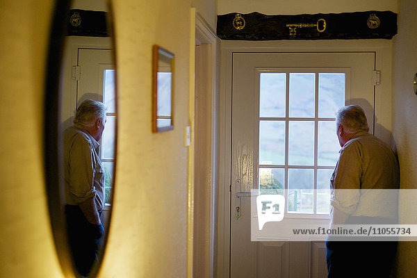 Ein älterer Mann  der an einer Tür steht und durch die Fensterscheiben nach draußen schaut.