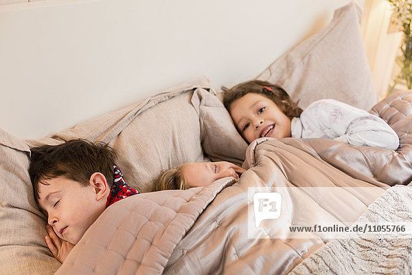 Three children lying in a bed under the covers.