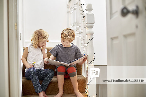Zwei Kinder  die zu Hause auf der Treppe nebeneinander sitzen und gemeinsam ein Buch lesen.