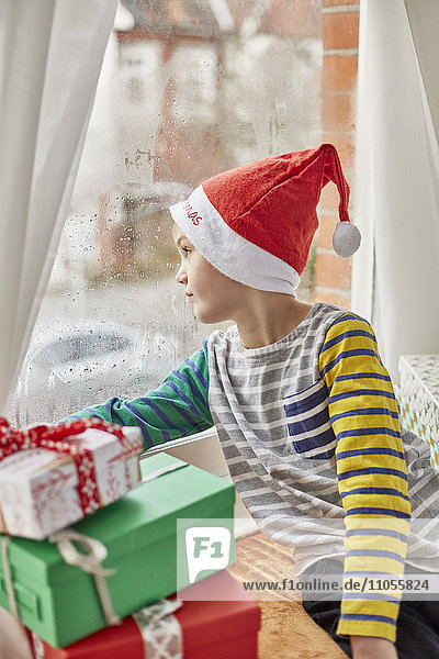 Weihnachtsmorgen in einem Familienhaus. Ein Junge mit Weihnachtsmannmütze,  der aus einem Schlafzimmerfenster schaut.