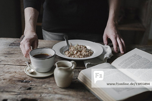 A person at a table with a cup of coffee  bowl of muesli and an open book.