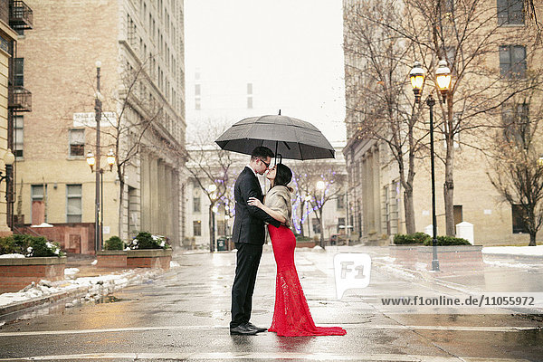 Eine Frau in einem langen roten Abendkleid mit Fischschwanzrock und Pelzstola und ein Mann in einem Anzug  die sich in der Stadt unter einem Regenschirm küssen.