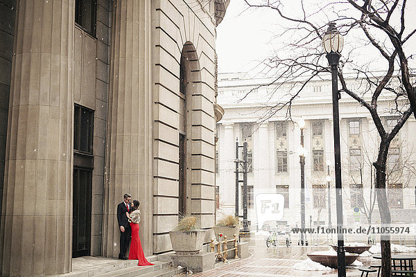 Eine Frau in einem langen roten Abendkleid mit Fischschwanzrock und einer Pelzstola und ein Mann im Anzug  der sich küsst  treten vor ein hohes Gebäude in einer Stadt.