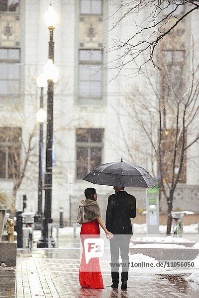 Eine Frau in einem langen roten Abendkleid mit Fischschwanzrock und Pelzstola und ein Mann in einem Anzug  die durch den Schnee in der Stadt spazieren.