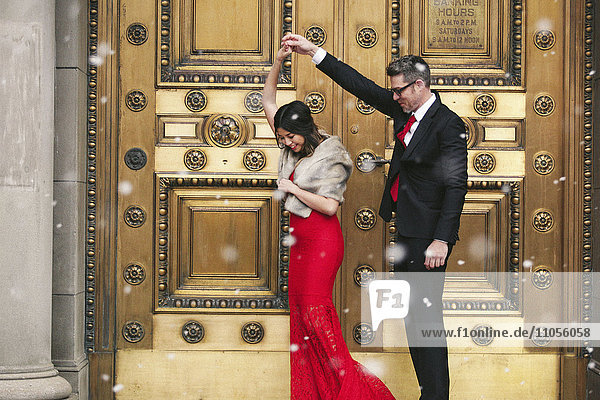 A woman in a long red evening dress with fishtail skirt and a fur stole  and a man in a suit dancing on the steps of a building.