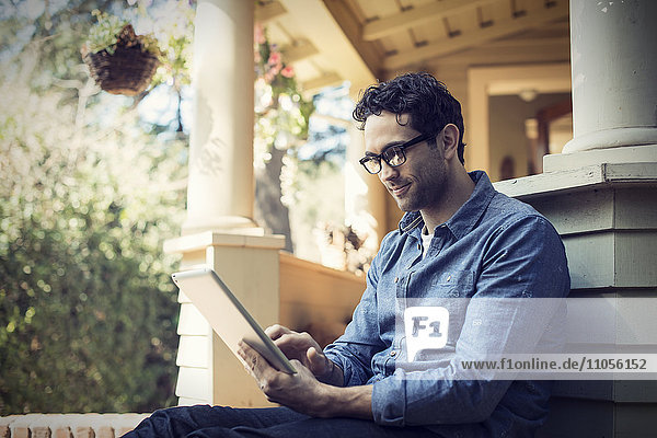 A man sitting relaxing in a quiet corner of a porch  using a digital tablet.