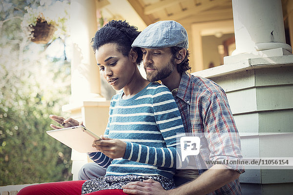 A couple  a man and woman seated on the porch steps  laughing  sharing a digital tablet.