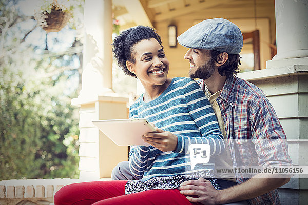 A couple  a man and woman seated on the porch steps  laughing  sharing a digital tablet.