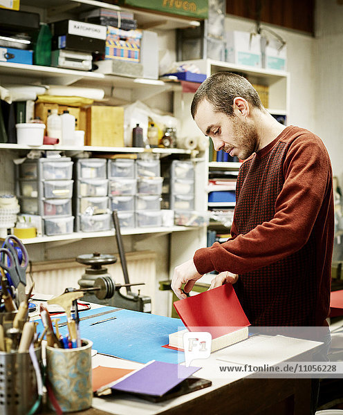 A man working in a book binding workshop  creating a red cover for freshly stitched pages.
