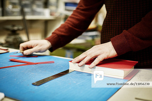 Ein Mann  der in einer Buchbinderei auf einer Werkbank arbeitet  um das Material für die Wiedergewinnung eines Buches zu messen und zu schneiden.
