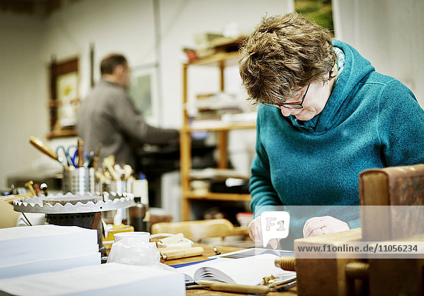 Eine Frau arbeitet an einem digitalen Tablett  umgeben von Buchbindewerkzeugen und -geräten.