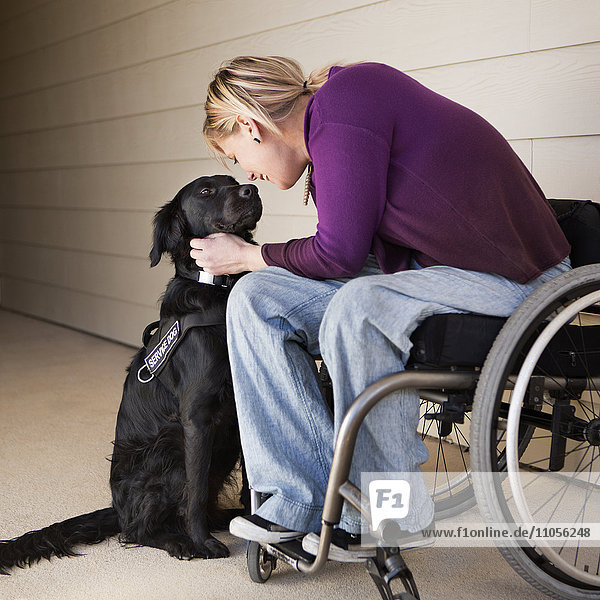 Eine reife Rollstuhlfahrerin streichelt ihren schwarzen Labrador-Diensthund und nimmt Augenkontakt mit dem Hund auf.