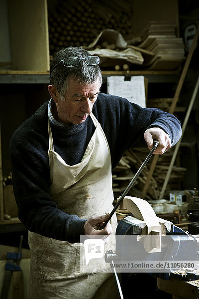 Ein Mann  der in der Werkstatt eines Möbelherstellers arbeitet und eine Raspel an einer Holzform in einer Klemme benutzt.