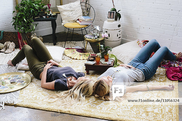 Zwei Frauen liegen auf dem Boden eines mit Kissen und persönlichen Gegenständen übersäten Raumes.