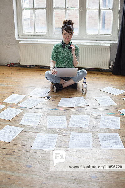 Junge Frau  die in einem Probenstudio mit einem Laptop auf dem Boden sitzt und sich Notenblätter ansieht.