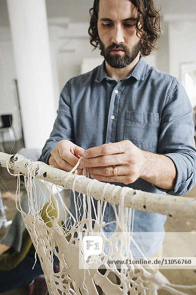 Ein Künstler arbeitet an einem Kunstwerk  das an einem Rahmen hängt und Fäden knüpft und webt.