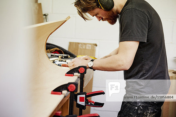 Eine Möbelwerkstatt,  die maßgeschneiderte zeitgenössische Möbelstücke unter Verwendung traditioneller Fertigkeiten im modernen Design herstellt. Ein Mann an einer Werkbank,  der an einem mit Klammern gehaltenen Stück gebogenen Holzes arbeitet.