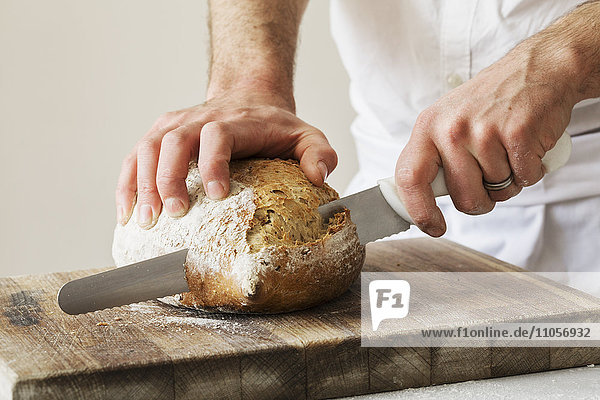 Nahaufnahme eines Bäckers  der mit einem Brotmesser einen frisch gebackenen Laib Brot schneidet.