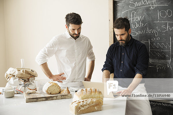 Zwei Bäcker  die mit einem Laptop an einem Tisch stehen  frisch gebackenes Brot  eine Tafel an der Wand.