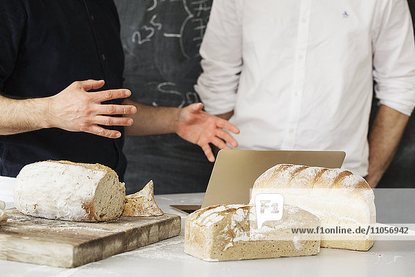 Close up of two bakers standing at a table  using a laptop computer  freshly baked bread.