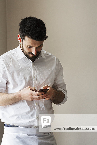 Halblanges Porträt eines bärtigen Mannes mit einer weißen Schürze  der sein Smartphone benutzt.