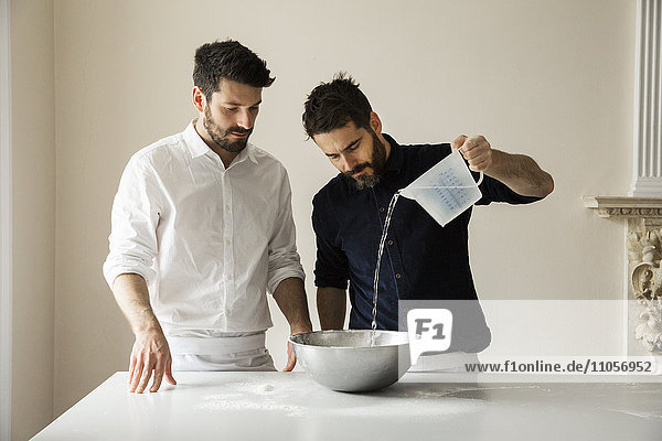 Zwei Bäcker stehen an einem Tisch  bereiten Brotteig zu  gießen Wasser aus einem Messbecher in eine Rührschüssel aus Metall.