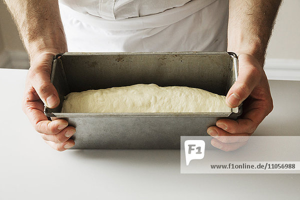 Nahaufnahme eines Bäckers  der eine mit Brotteig gefüllte Backform hält.