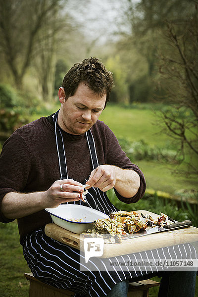 Chef sitting outdoors with a chopping board on his lap  preparing a crab.