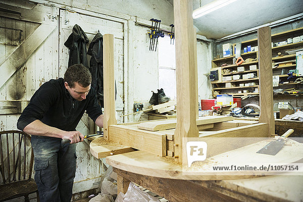 Ein Mann in einer Schreinerei  der mit einem Hammer an der Kante eines neuen Holztisches arbeitet.