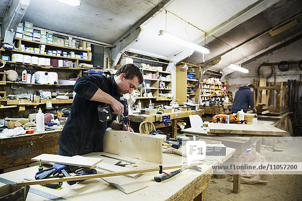 Man standing at a work bench in a carpentry workshop  working on a piece of wood  using a drill.