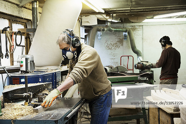 Man standing in a carpentry workshop  wearing a respirator and hearing protector  working on a piece of wood.