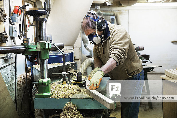 Mann steht in einer Schreinerei  trägt ein Atemschutzgerät und Gehörschutz und arbeitet an einem Stück Holz.