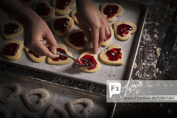 Valentinstag beim Backen,  Frau streicht Himbeermarmelade auf herzförmige Kekse.