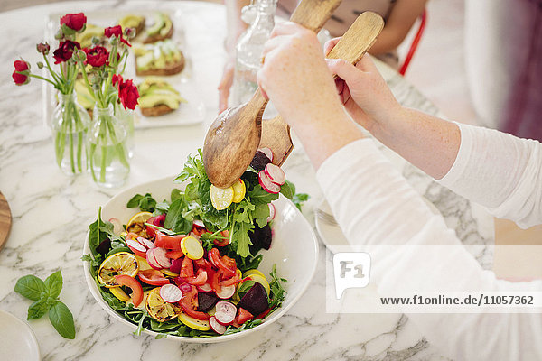 Blick von oben auf einen mit Besteck und Tellern mit zubereiteten Speisen gedeckten Tisch. Eine Frau nimmt eine Portion Salat.
