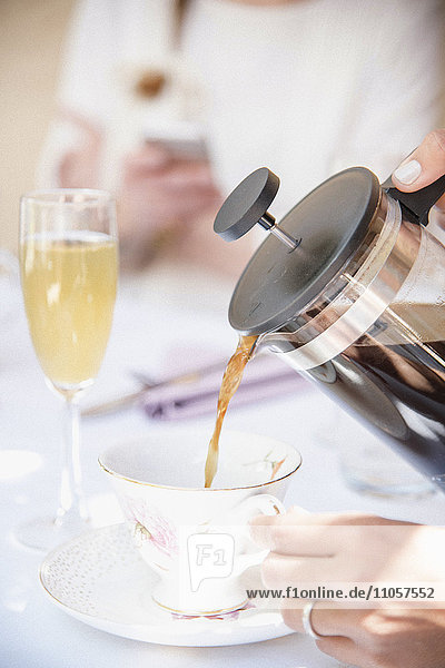 Nahaufnahme eines Kaffees,  der aus einer französischen Kaffeepresse in eine Tasse,  ein Glas Champagner,  gegossen wird.