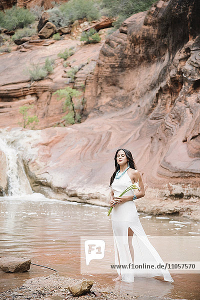 Junge Frau mit langen braunen Haaren  trägt ein langes weißes Kleid  steht an einem Fluss und hält Aronstab-Lilien.