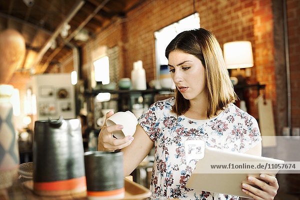 Junge Frau in einem Geschäft  die ein digitales Tablett und einen Keramikbecher in der Hand hält.
