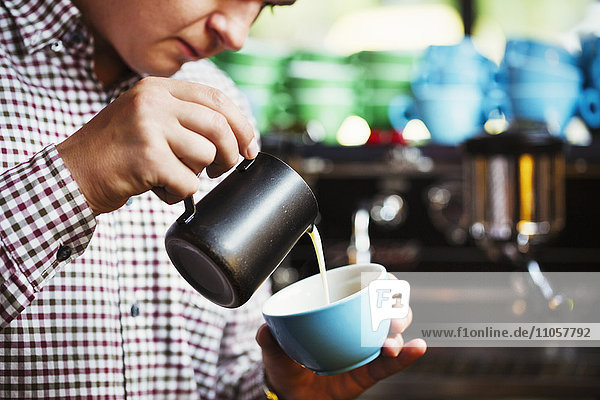 Spezialisiertes Kaffeehaus. Ein Mann gießt heiße Milch in eine Tasse Kaffee,  um ein Muster auf der Oberseite zu machen.