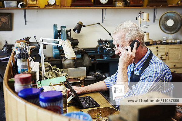 Ein Uhrmacher  der in seiner Werkstatt beschäftigt ist  mit seinem Laptop und Festnetztelefon.