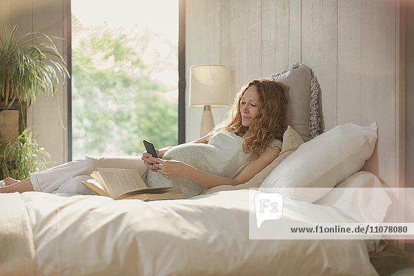 Pregnant woman laying relaxing with book and texting with cell phone in bed