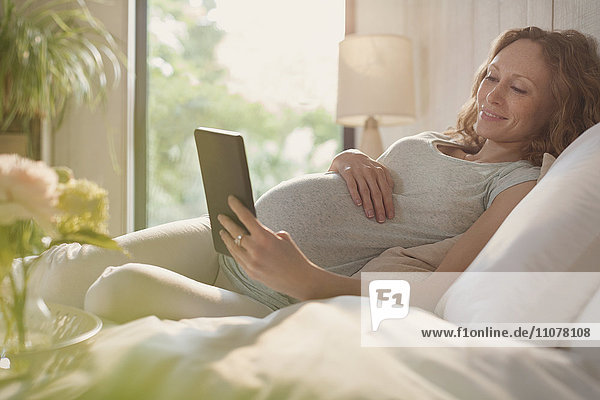 Schwangere Frau entspannt sich mit digitalem Tablett im sonnigen Schlafzimmer