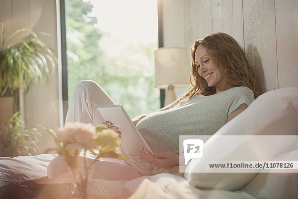 Lächelnde Schwangere mit digitalem Tablett auf dem Bett im sonnigen Schlafzimmer