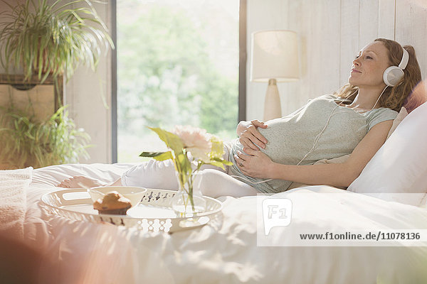 Serene pregnant woman listening to music with headphones on bed