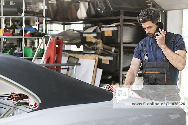 Mechaniker im Gespräch mit dem Handy beim Bestellen von Autoteilen aus dem Katalog in der Kfz-Werkstatt