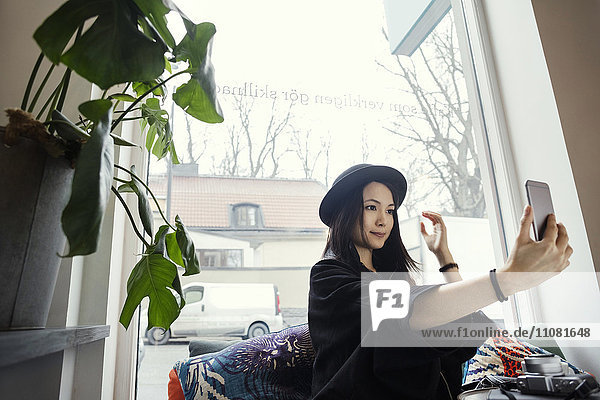 Lächelnde junge Frau nimmt Selfie  während sie im Büro am Fenster sitzt.