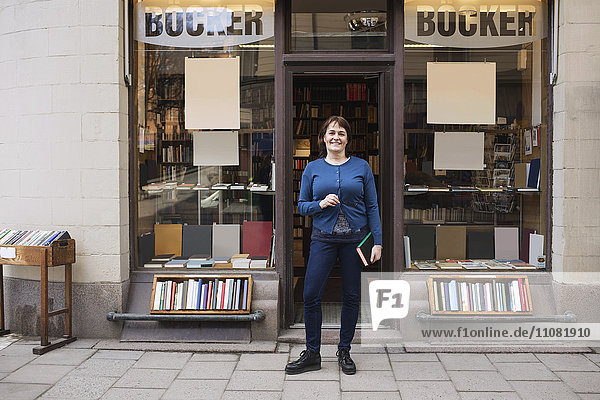 Portrait of happy woman holding book while standing outside bookstore